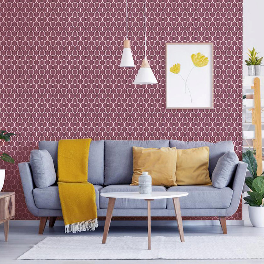 A modern living room with a gray sofa, yellow pillows, and a yellow throw blanket. There’s a white round coffee table, two pendant lights, a floral wall art, and a maroon hexagon-tiled accent wall. Plants and shelves complete the decor.