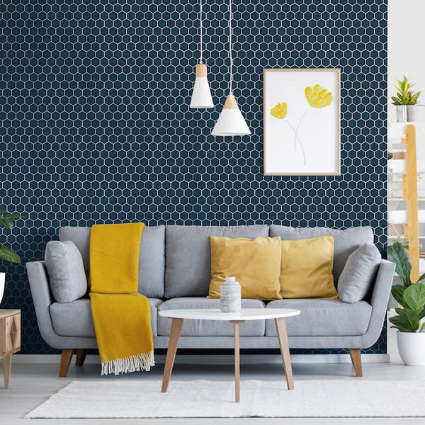 A modern living room with a grey sofa, yellow cushions and throw, round white coffee table, pendant lights, and a dark hexagonal patterned accent wall with a framed yellow floral print. Plants and shelves decorate the space.