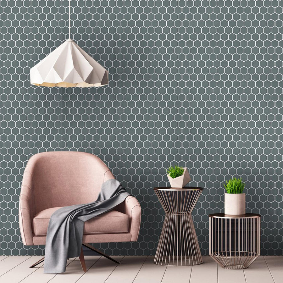 A light pink modern armchair with a gray throw is next to two wire-frame side tables holding potted plants. A geometric white pendant lamp hangs above, and the wall features a dark hexagon tile pattern.