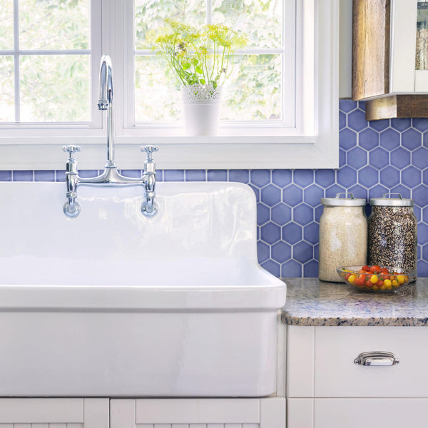 A white farmhouse sink beneath a window with green plants outside, featuring a chrome faucet, blue hexagon tile backsplash, two jars with grains, and a bowl of tomatoes on a speckled countertop.