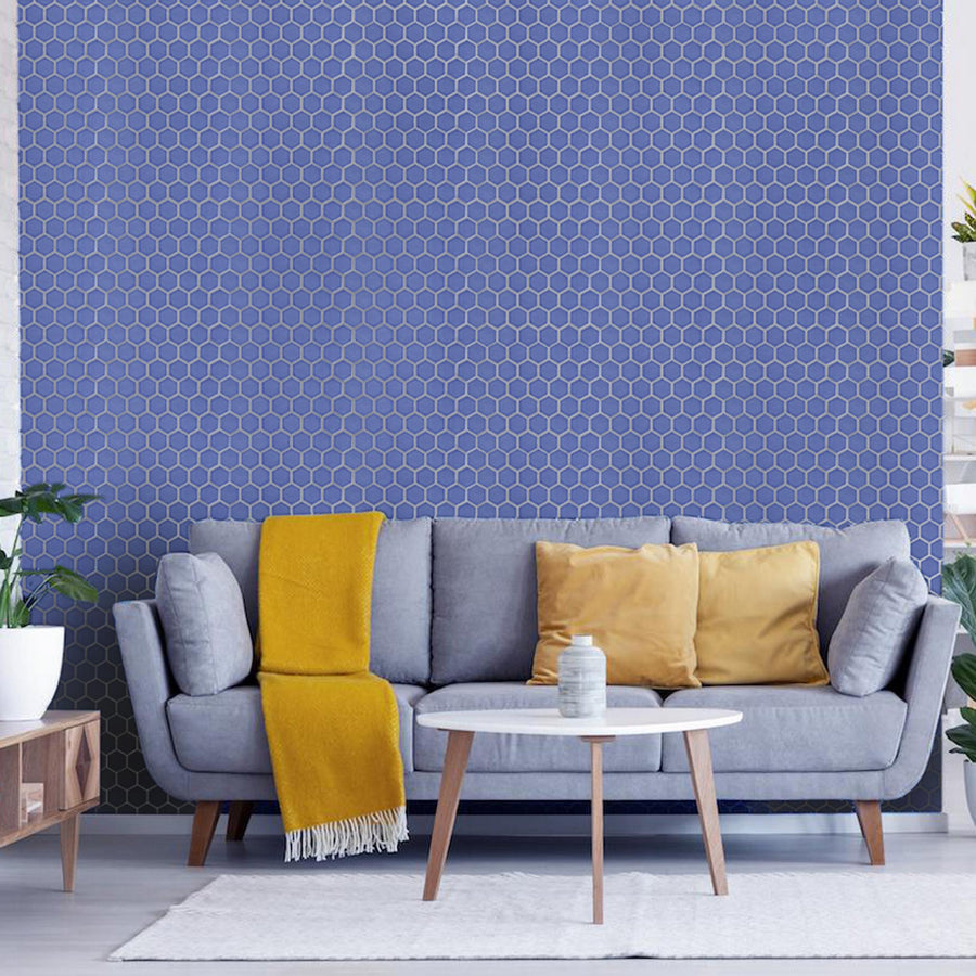A modern living room with a grey sofa, yellow throw blanket, and yellow cushions. A small white round coffee table is in front, with green plants and shelves visible. The background wall has a blue hexagonal tile pattern.
