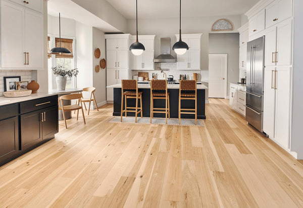 Spacious modern kitchen with light wood floors, white cabinets, black island with wicker barstools, pendant lights, stainless steel appliances, and a small dining area with two wooden chairs and a table.