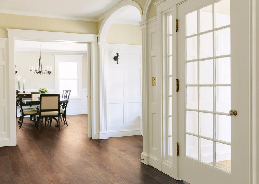 Bright entryway with glass-paned door, white walls, and wood flooring leading to a dining room with a table, chairs, and a chandelier in the background. Natural light fills the elegant, open space.