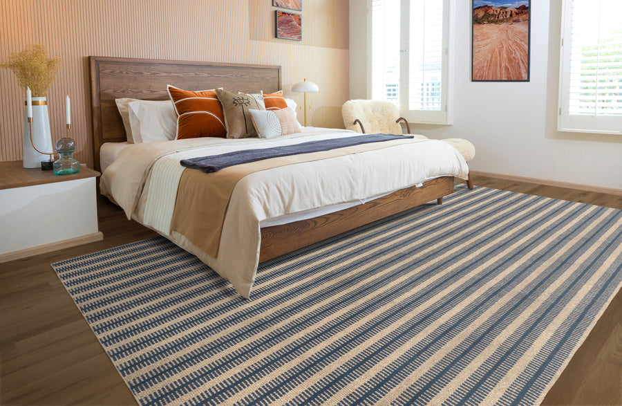 A modern bedroom with a wooden bed, white and tan bedding, orange and beige pillows, a striped blue and beige rug, white nightstand, and large windows letting in natural light.