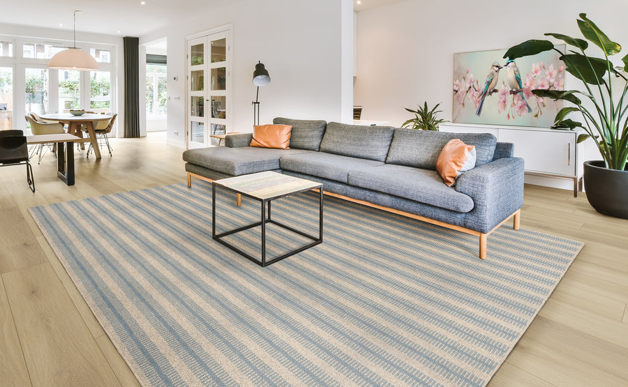 A modern living room with a gray sectional sofa, two orange pillows, a striped area rug, a square coffee table, large potted plants, wall art with pink flowers, and dining area visible in the background.