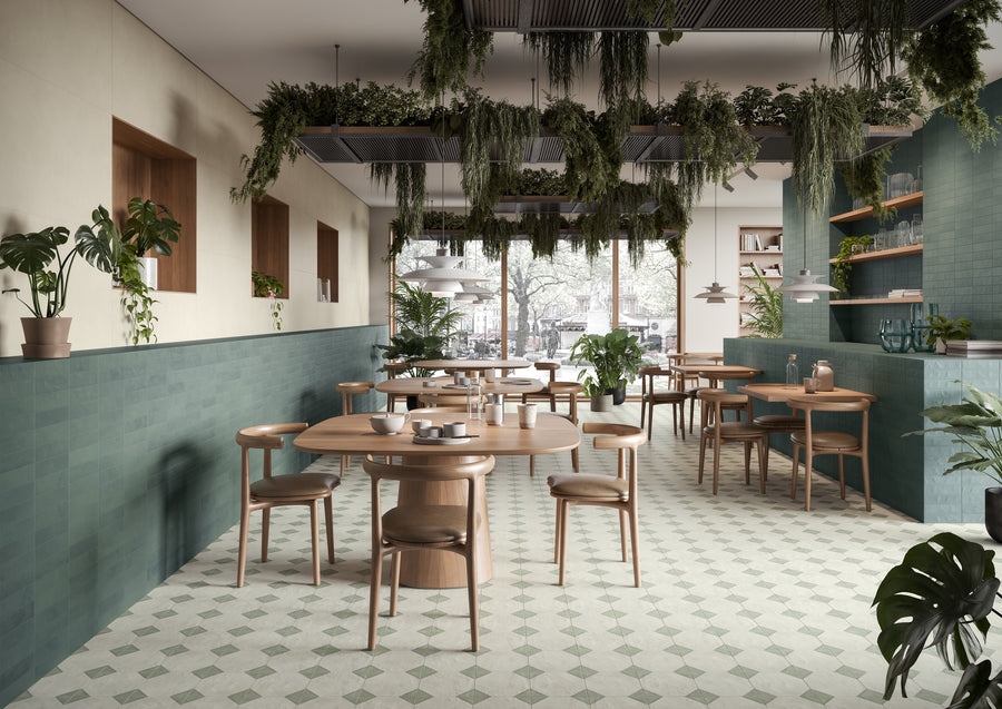 A modern, airy café with wooden tables and chairs, green tiled walls, potted plants, and lush greenery hanging from the ceiling. Large windows let in natural light, creating a cozy and inviting atmosphere.