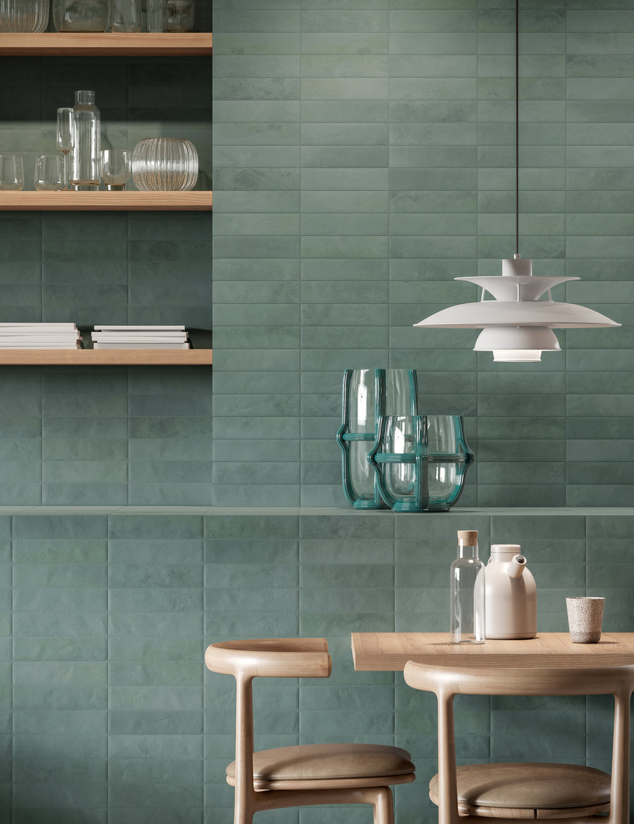 A modern dining area with green tiled walls, wooden shelves displaying glassware, a wooden table with chairs, clear and white vases, and a white pendant lamp hanging above.