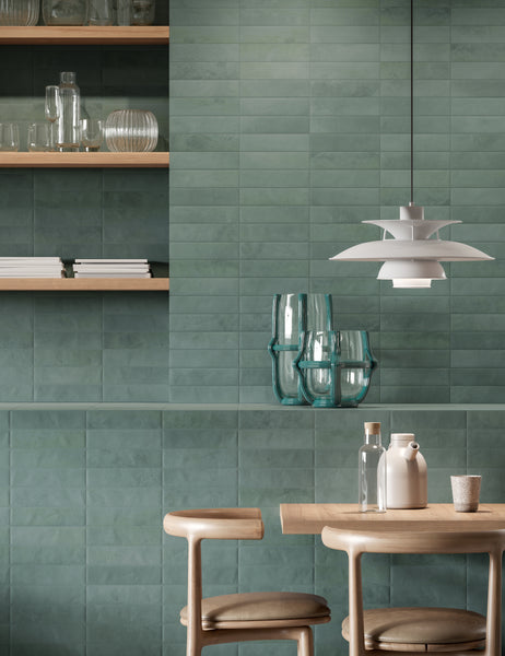 A modern dining area with green tiled walls, wooden shelves displaying glassware, a wooden table with chairs, clear and white vases, and a white pendant lamp hanging above.