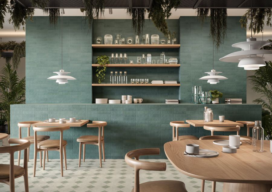 Modern cafe with light wood tables and chairs, green tiled walls, open wooden shelves holding dishes and glassware, white pendant lights, and plants hanging from the ceiling and placed around the space.