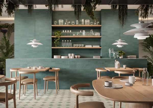 Modern cafe with light wood tables and chairs, green tiled walls, open wooden shelves holding dishes and glassware, white pendant lights, and plants hanging from the ceiling and placed around the space.