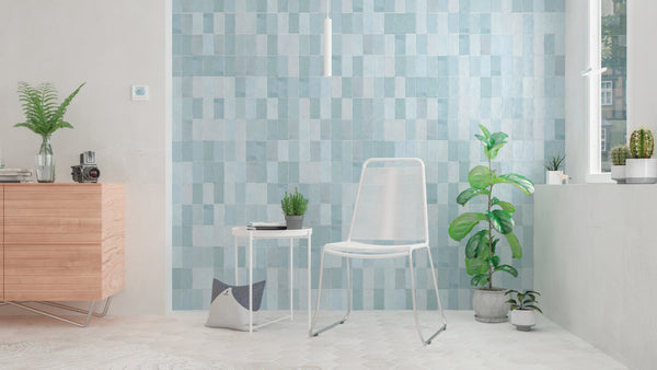 Minimalist room with light blue square-tiled wall, white metal chair, small white side table with potted cactus, green plants, wooden cabinet, camera, and sunlight streaming through a window.
