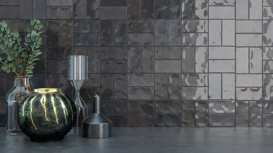 A decorative black and gold vase, glass bottles, an hourglass, and a metal funnel sit on a gray surface in front of a glossy, dark tiled wall with a patchwork pattern.