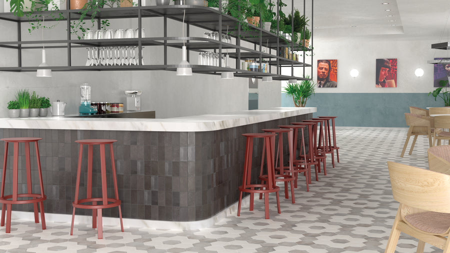 A modern bar with a marble countertop, red bar stools, hanging glassware, and potted plants. There are geometric floor tiles, artwork on the walls, and wooden chairs in the background. The space is bright and contemporary.