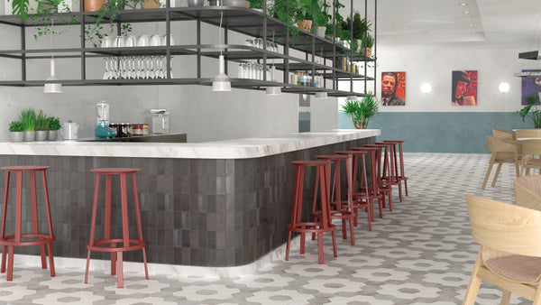 A modern bar with a marble countertop, red bar stools, hanging glassware, and potted plants. There are geometric floor tiles, artwork on the walls, and wooden chairs in the background. The space is bright and contemporary.