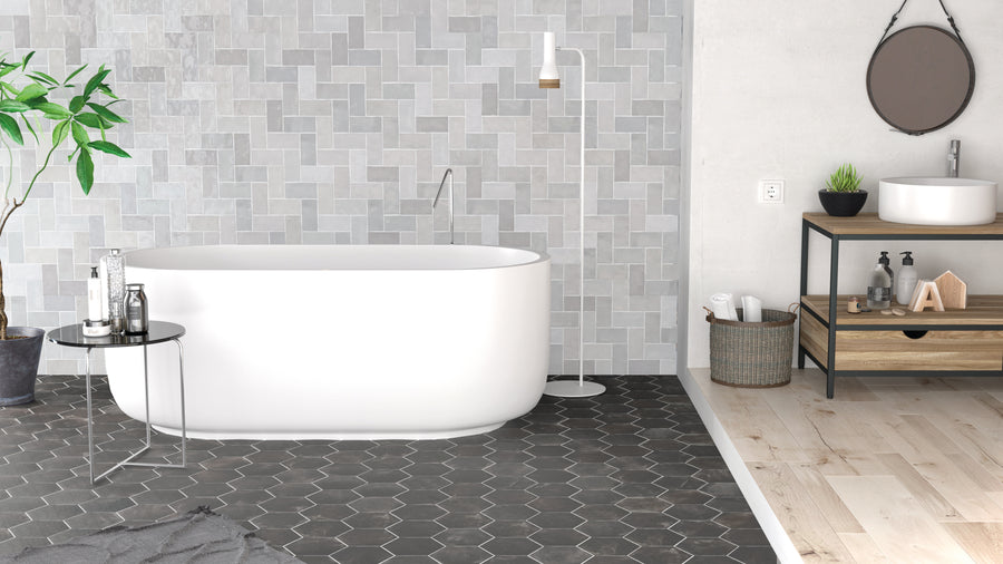 Modern bathroom with a white freestanding tub, gray geometric tile wall, hexagonal black floor tiles, a small table, potted plant, round mirror, vessel sink on a wooden stand, and a basket under the sink.