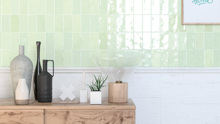 A wooden sideboard holds decorative items, including vases, a candle, a plant in a white pot, and a glass lamp. The background features glossy green tiles and a partial framed artwork on the wall.