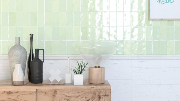 A wooden sideboard holds decorative items, including vases, a candle, a plant in a white pot, and a glass lamp. The background features glossy green tiles and a partial framed artwork on the wall.