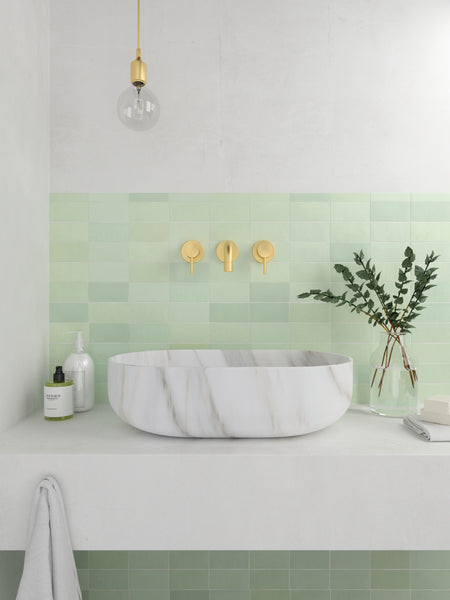 A modern bathroom sink with a white marble basin, gold fixtures, and light green square tiles on the wall. There is a glass vase with greenery, a soap dispenser, and a towel hanging below the counter.