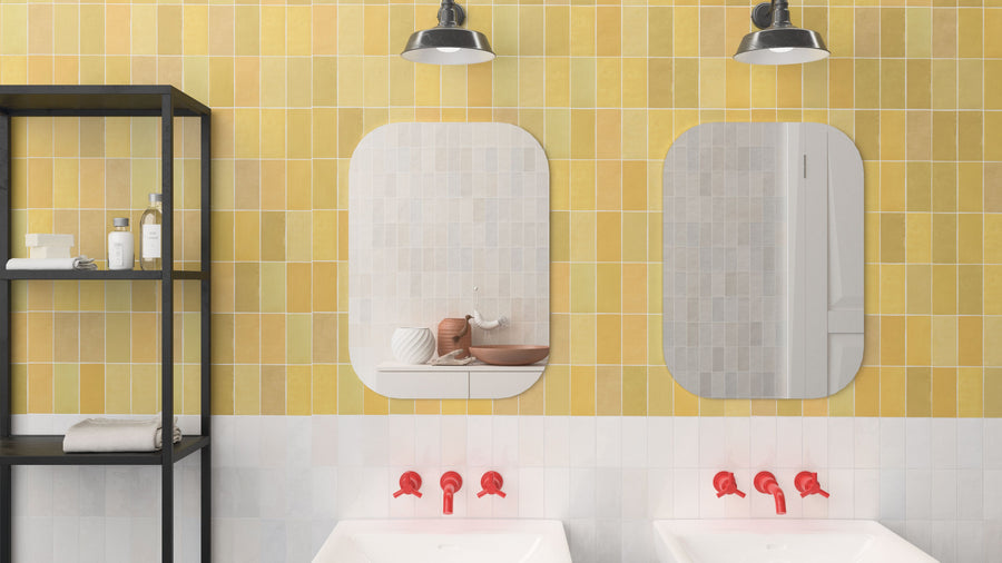 A bathroom with yellow and white tiled walls, two mirrors above double sinks with red faucets, black shelving on the left holding towels and toiletries, and two black industrial lights above the mirrors.