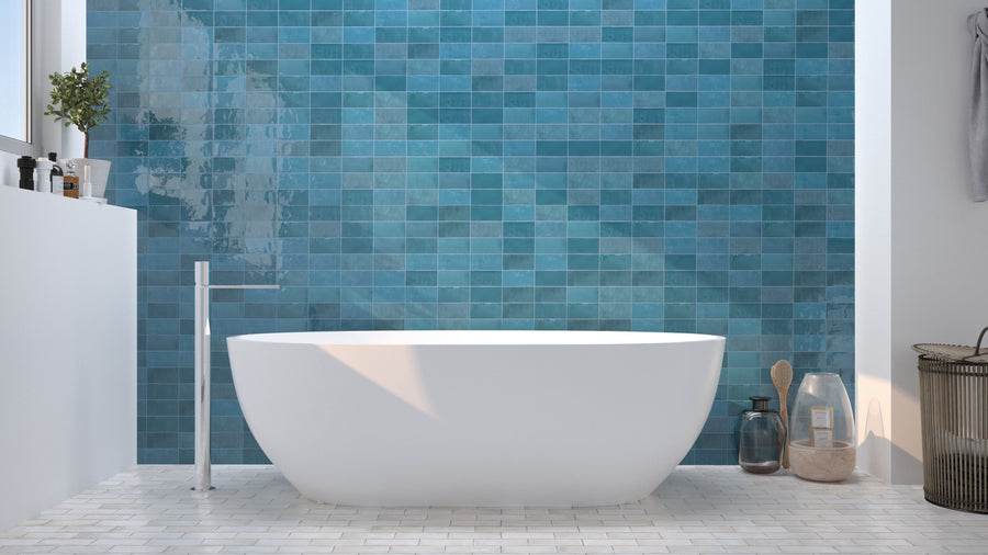 Modern bathroom with a white freestanding tub, blue square-tiled wall, white floor tiles, a tall faucet, and various bath accessories on the floor and shelf, with natural light from a window.