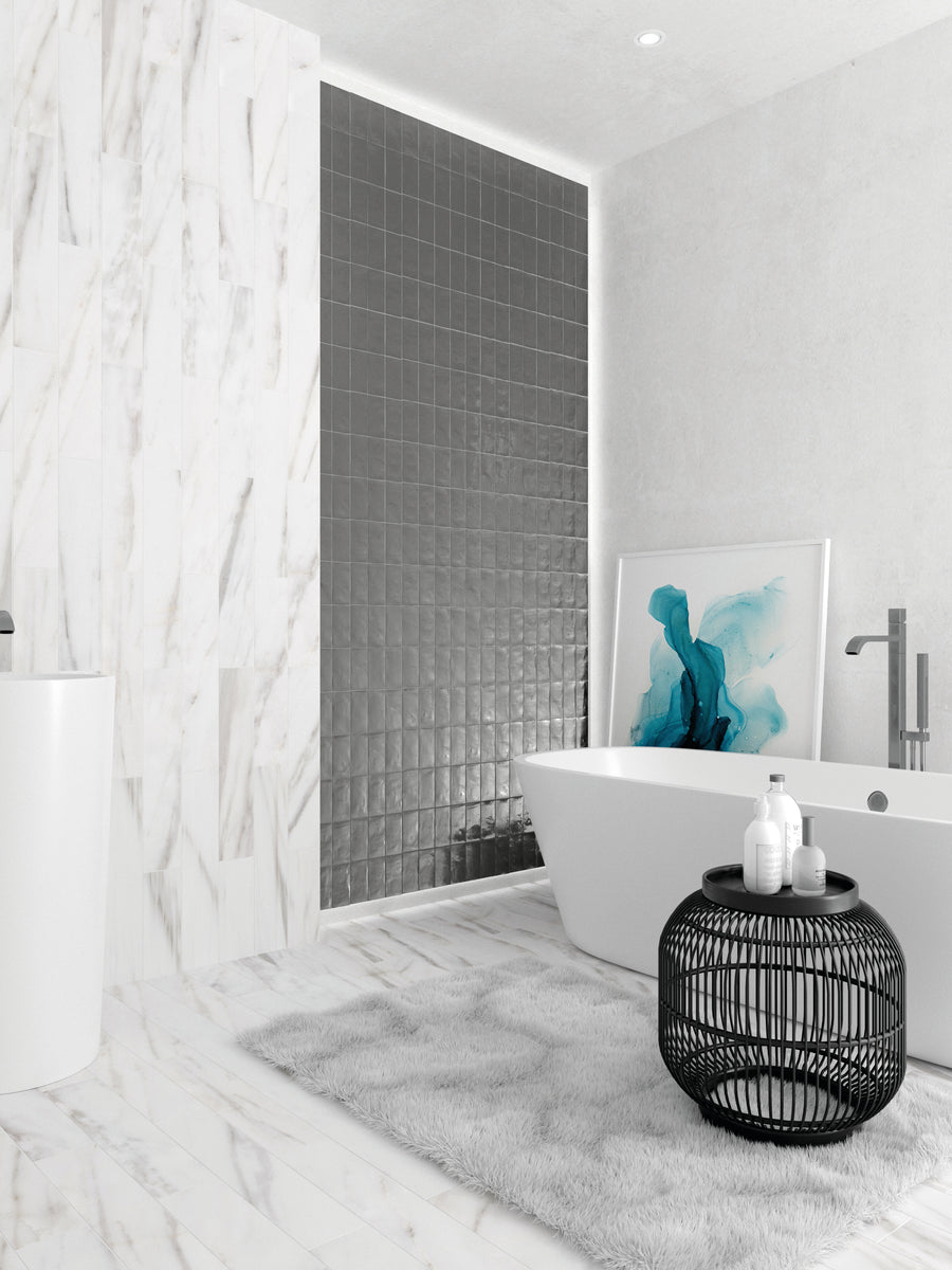 Modern bathroom with marble walls and floor, a freestanding white bathtub, black tiled accent wall, round black wire table with toiletries, gray rug, and blue abstract artwork resting beside the tub.