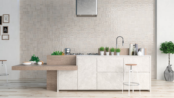 Modern kitchen with a white and beige color scheme, featuring a minimalist island with potted herbs, bar stools, a built-in stove, and a textured tile backsplash. Decor includes framed art and a potted plant.