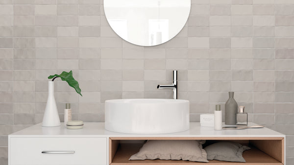 A modern bathroom vanity with a round white sink, chrome faucet, decorative vases, and toiletries. A round mirror hangs above, and two pillows are stored in an open shelf below the counter. The wall is tiled in light gray.