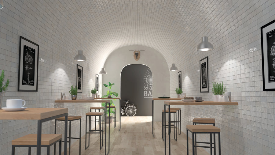 A modern, minimalist café with white tiled walls, wooden tables, and black stools. Green plants decorate the tables. A bicycle and wall art are visible at the far end under a curved ceiling.