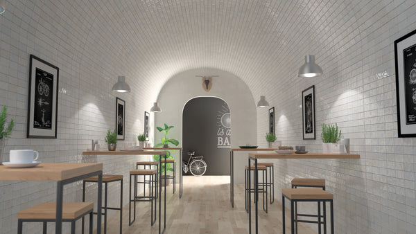 A modern, minimalist café with white tiled walls, wooden tables, and black stools. Green plants decorate the tables. A bicycle and wall art are visible at the far end under a curved ceiling.