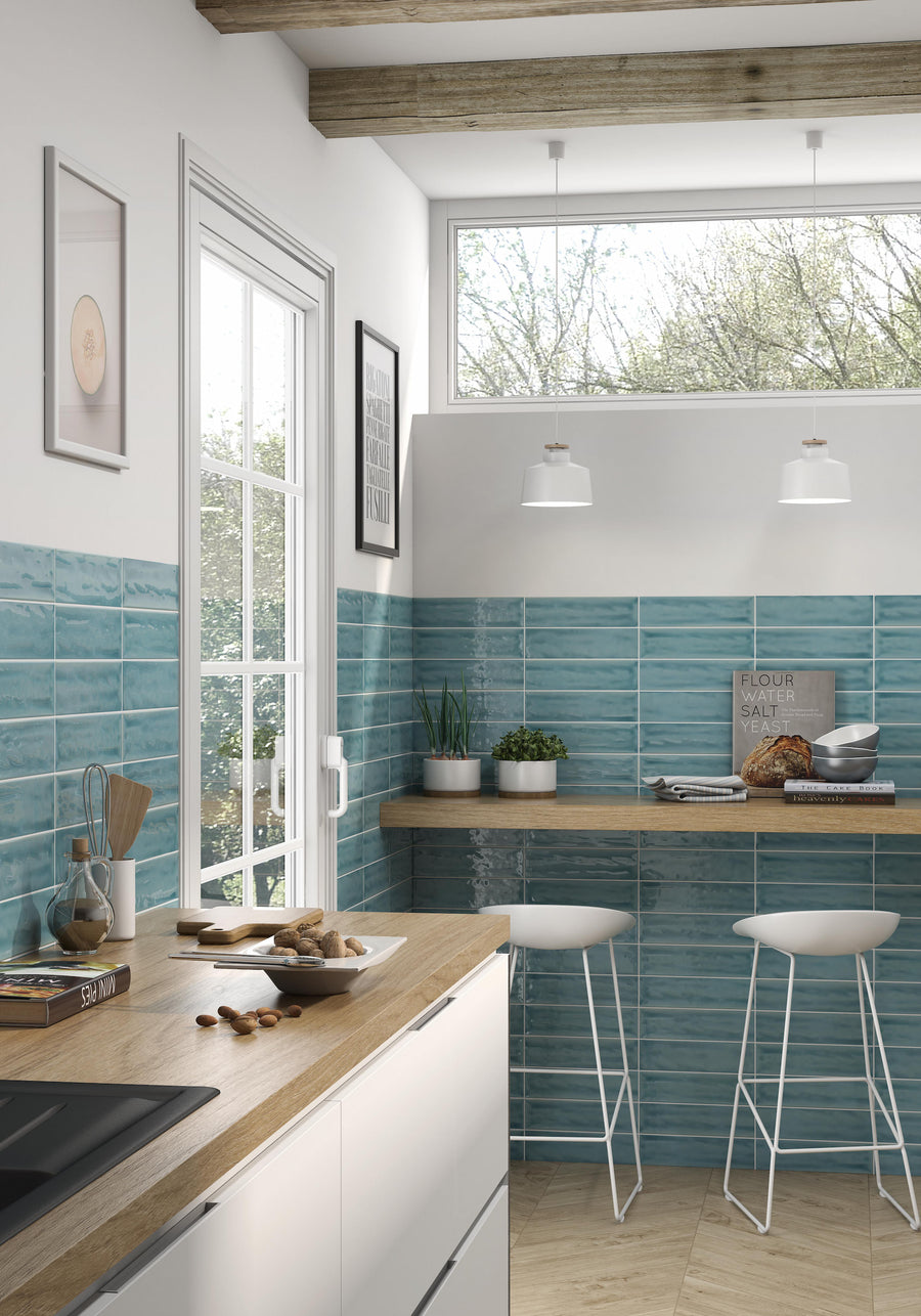 Modern kitchen with teal subway tile backsplash, wooden countertops, and white cabinets. Two white bar stools sit at a breakfast bar by large windows with a view of trees. Decor includes framed art and potted plants.