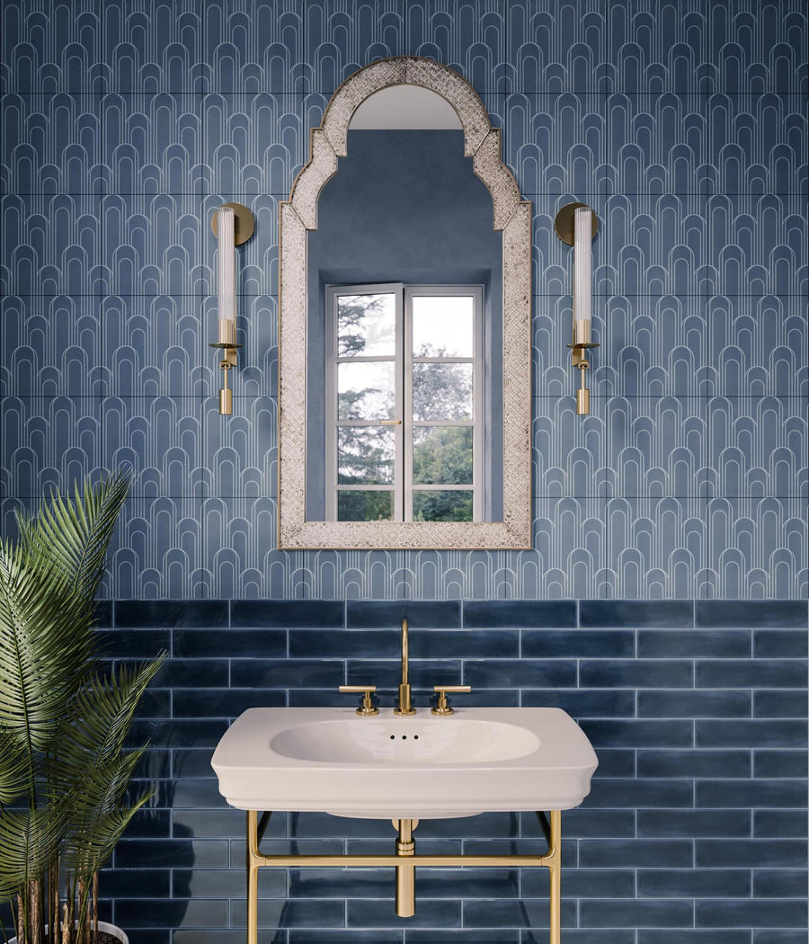 A bathroom with blue patterned tile walls, a white sink with gold fixtures, a gold-framed arched mirror, two wall sconces, and a potted plant on the left. A window is visible in the mirror’s reflection.