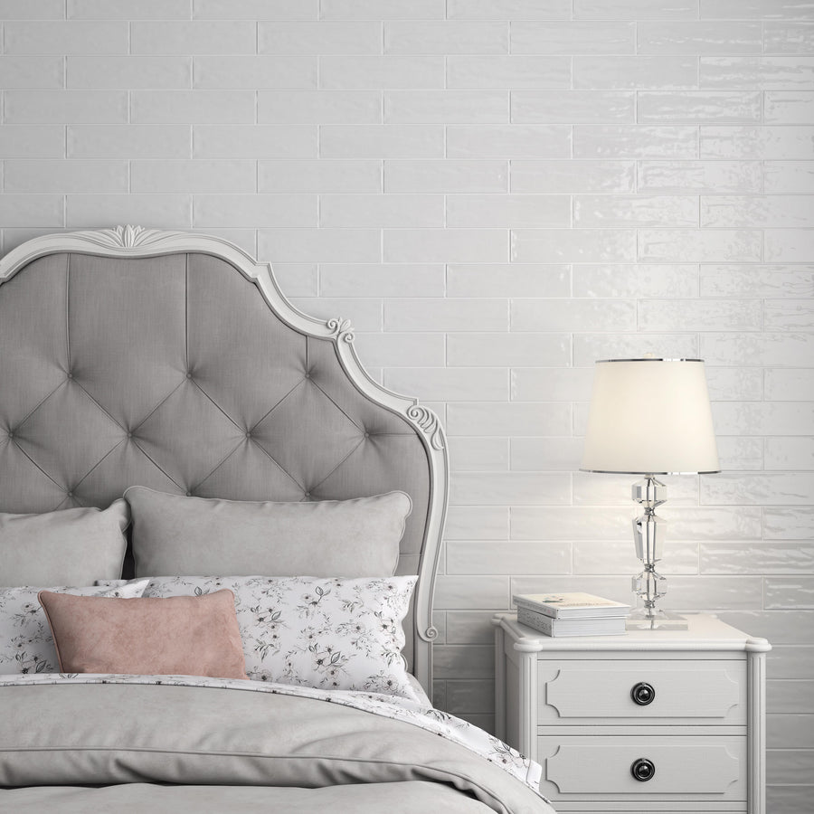 A stylish bedroom with a tufted gray headboard, layered bedding in gray and floral patterns, a small pink pillow, and a white nightstand holding books and a glass lamp against a white brick wall.