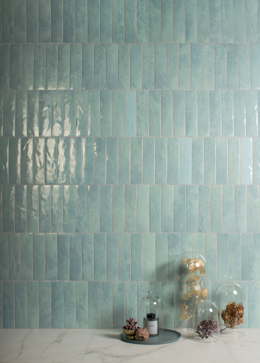 Glossy, rectangular aqua tiles cover a wall in a vertical pattern. On a white marble surface below, three glass cloches display dried flowers and succulents, arranged in a minimalist, decorative style.