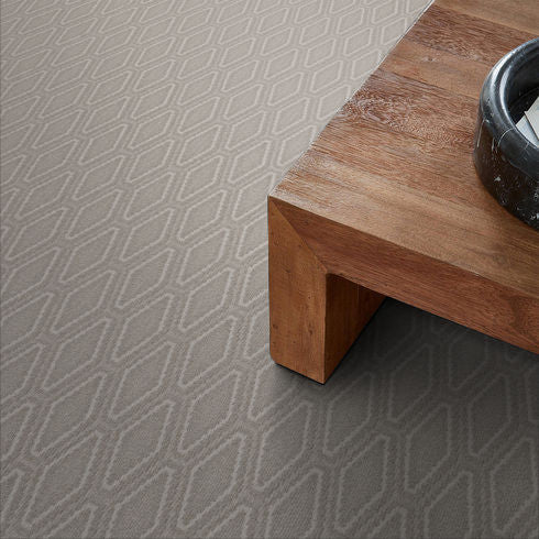 A close-up view of a geometric-patterned gray carpet next to a corner of a wooden table with a black circular object on top.