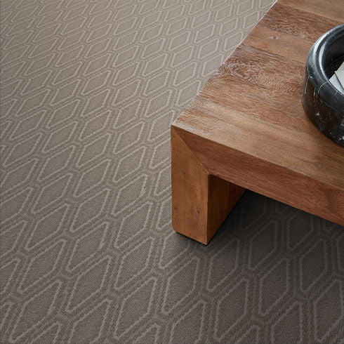 A close-up view of a geometric-patterned beige carpet next to a wooden table with a black bowl on top.