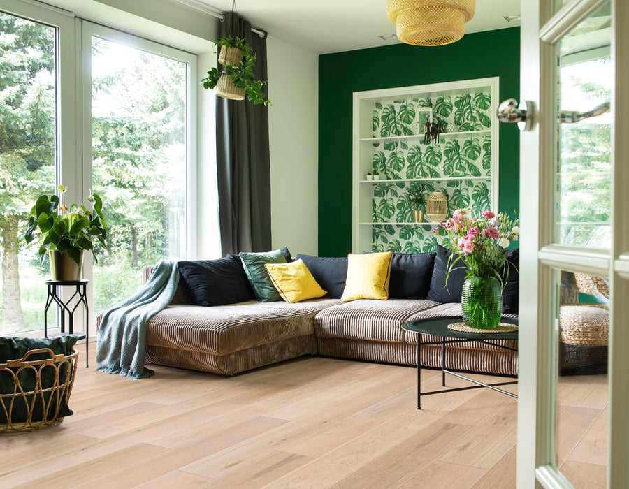 A cozy living room with a large sectional sofa, colorful pillows, a round coffee table with a vase of flowers, potted plants, and large windows overlooking greenery; a green accent wall features tropical wallpaper.