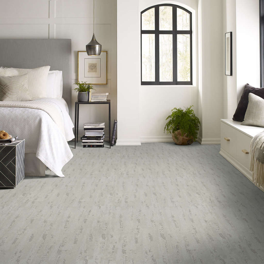 A modern, bright bedroom with a large bed, light gray carpet, gray headboard, side table with lamp and decor, a window seat, arched window, and a potted plant on the floor.