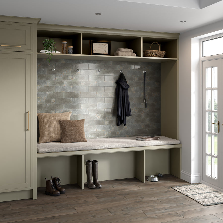A mudroom with a built-in bench, beige cushions, brown pillows, coat hooks with jackets, cubby storage above, boots on the floor, a dog bowl, and a door leading outside. Gray tiled wall and wood flooring complete the space.