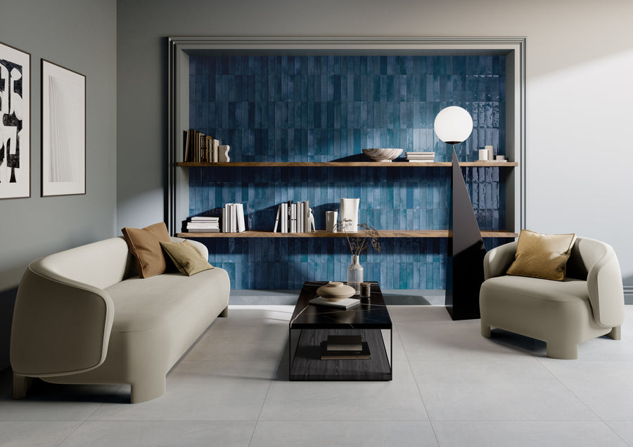 Modern living room with light sofas, a black coffee table, and a built-in blue-tiled bookshelf wall. Shelves hold books and decor items; a geometric lamp stands on the right. Minimalist art hangs on the left wall.