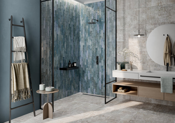 Modern bathroom with a blue-green tiled shower, glass partition, and rainfall showerhead. Vanity with round mirror, open shelves, and decorative items. Ladder shelf holds towels, and a small side table has candles and a jar.