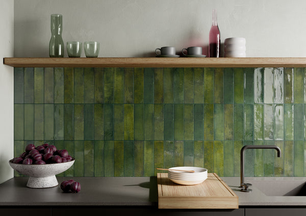 A kitchen counter with a bowl of plums, a wooden cutting board with a bowl and chopsticks, and a sleek faucet. Above, a shelf holds cups, glasses, and a bottle against a green tile backsplash.