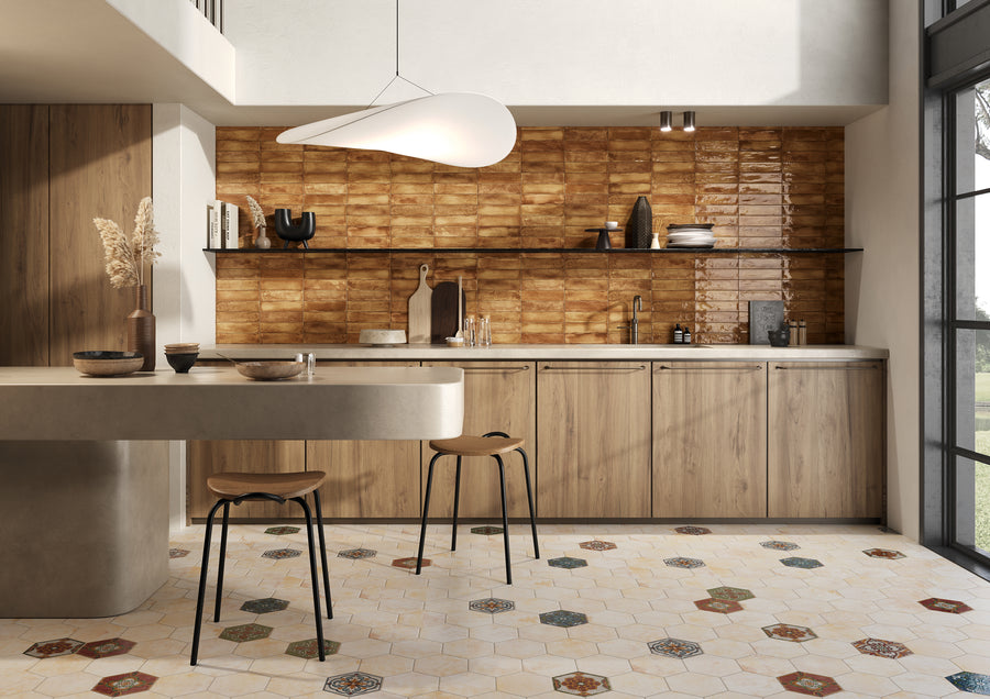 Modern kitchen with light wood cabinets, tan countertops, and a warm brown tile backsplash. Two wooden stools sit by a curved island. Patterned hexagonal floor tiles and large windows create an airy, bright atmosphere.