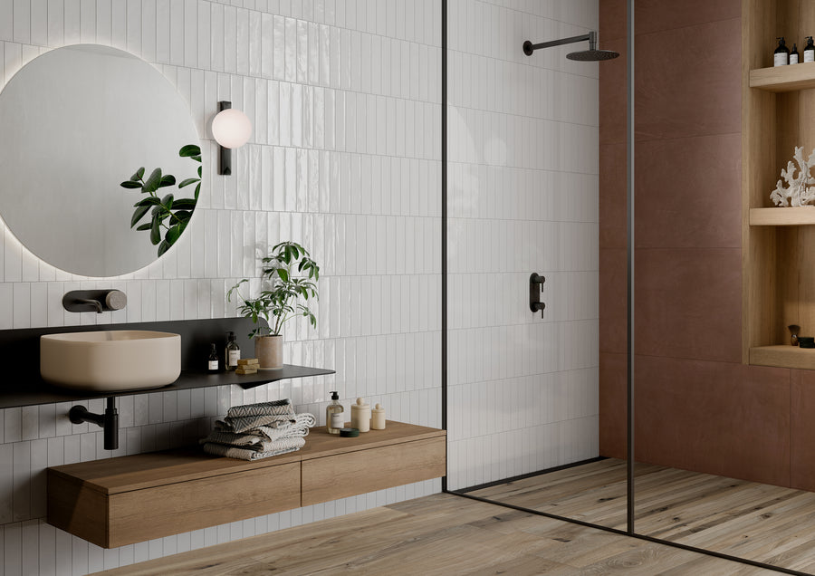 Modern bathroom with a floating wooden vanity, round mirror, wall sconce, potted plant, open shelving, and a glass-enclosed shower featuring minimalist fixtures. White tile and wood accents create a clean, contemporary look.