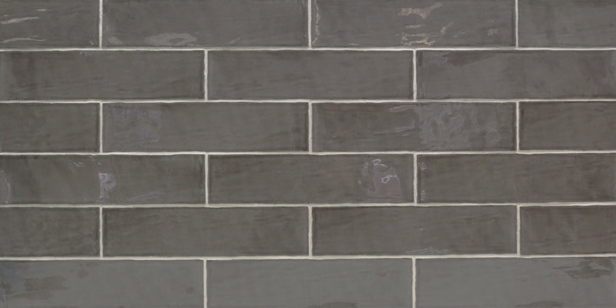 The Masia 3" x 12" Subway Tile in Gris Oscuro features a glossy dark gray finish and is arranged in a staggered brick pattern with light grout, making it perfect for accent walls or modern kitchen backsplashes.