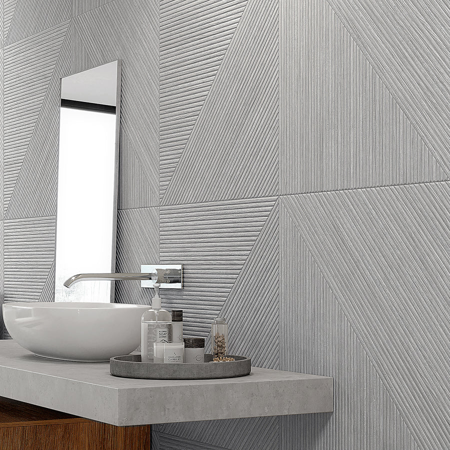 A modern bathroom with textured gray geometric wall tiles, a rectangular mirror, a round white sink, and a concrete countertop. Toiletries are arranged neatly on a tray beside the sink.