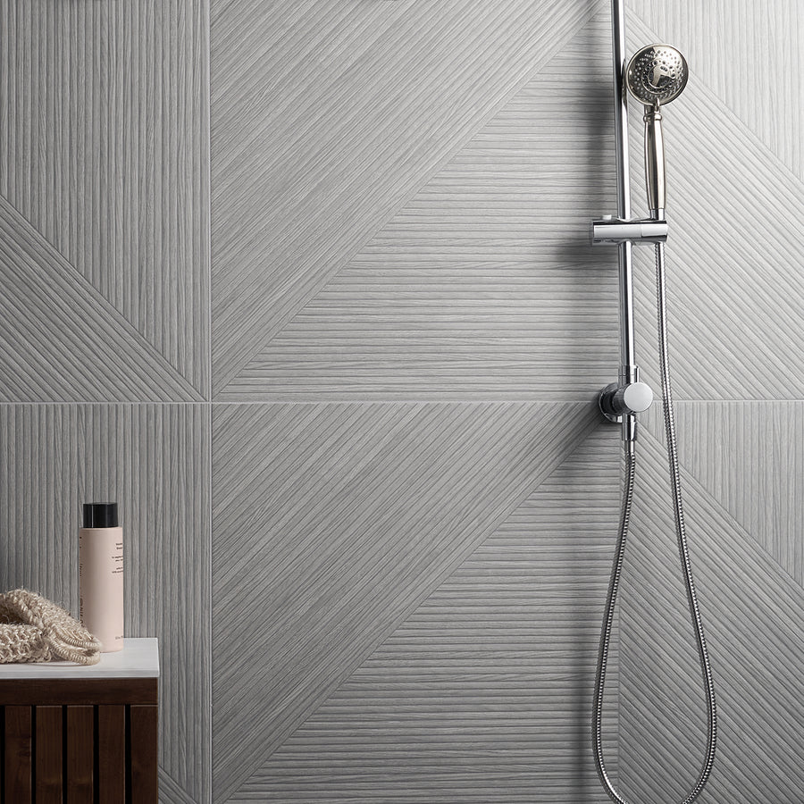 A modern shower with a handheld showerhead against light gray geometric patterned tiles. A wooden bench with a beige towel and a bottle stands to the left.