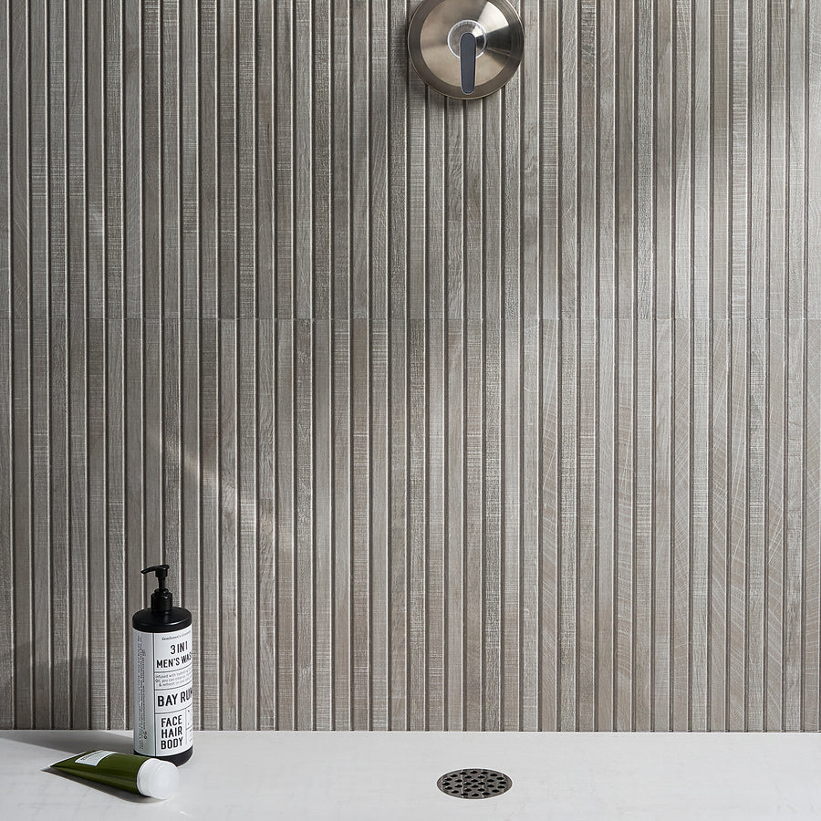 Modern shower with vertically textured gray wall tiles, a round metal shower control, a metal drain on the white shower floor, and two toiletries: a tube and a bottle labeled 3 in 1 Mens Wash.