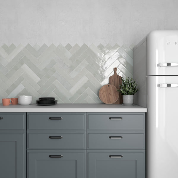 A modern kitchen with blue-gray cabinets, a white retro refrigerator, and a light herringbone tile backsplash. On the counter are dishes, cups, cutting boards, and a potted plant.