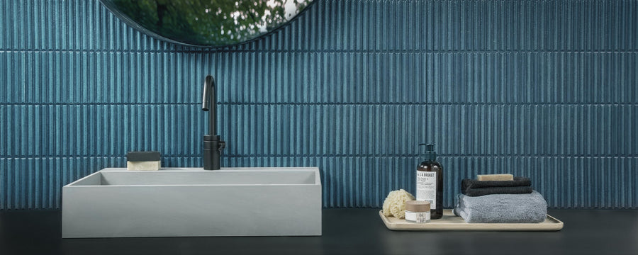 Modern bathroom counter with a rectangular gray sink, black faucet, bar soap, and a tray holding folded towels, a sponge, a jar, and a bottle, set against blue textured tile walls and a round mirror.