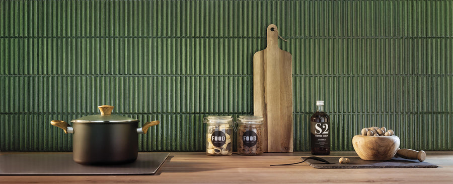 A modern kitchen countertop with a black pot, two jars labeled FOOD, a cutting board, a bottle of sauce, and a mortar and pestle, set against a textured green tile backsplash.